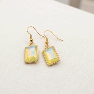 May include: A pair of gold-colored earrings with rectangular, opalescent-looking stones. The stones have a soft, yellow and blue iridescence. The earrings are on a white surface.