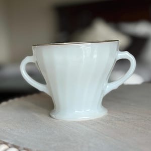 May include: A white milk glass sugar bowl featuring a subtle swirl pattern, two side handles, and a flared base. The rim of the bowl is finished with a thin gold metallic band, providing a simple contrast to the opaque white glass body.