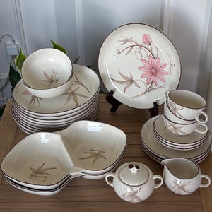 May include: A cream-colored ceramic dinnerware set featuring a brown rim and a floral pattern with pink and brown petals. The collection includes stacked plates, bowls, a divided serving dish, teacups with saucers, a lidded sugar bowl, and a creamer.