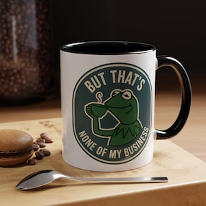 May include: White ceramic mug with a black interior and handle. The mug features a green and white graphic of Kermit the Frog holding a cup, with the text "BUT THAT'S NONE OF MY BUSINESS" in a circular design.