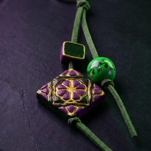 Peut inclure: Un porte-clés composé d'un cordon en daim vert attaché à un anneau en métal argenté. Le cordon retient un pendentif carré violet et doré à motif floral, une petite perle carrée vert foncé et une perle ronde vert vif avec des motifs floraux peints.
