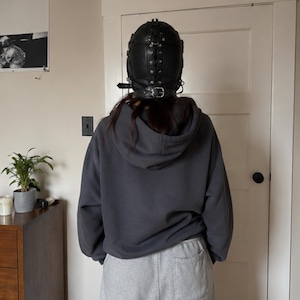 May include: A black leather headgear featuring a central lace-up closure with metal eyelets, side buckles, and a thick adjustable neck strap. The headgear is worn with a dark gray hooded sweatshirt and light gray sweatpants, viewed from the back.