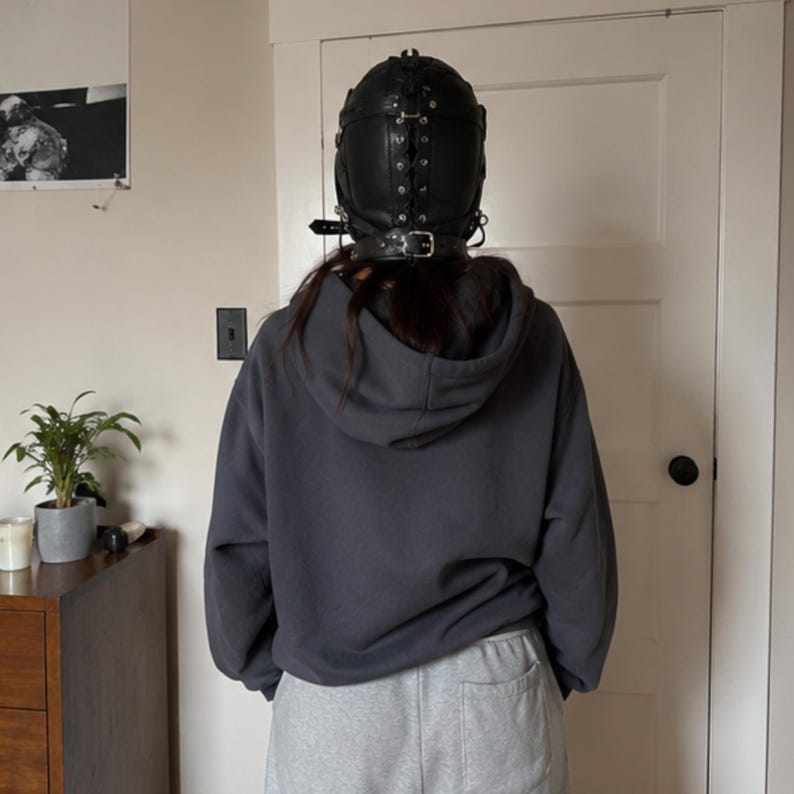 May include: A black leather headgear featuring a central lace-up closure with metal eyelets, side buckles, and a thick adjustable neck strap. The headgear is worn with a dark gray hooded sweatshirt and light gray sweatpants, viewed from the back.