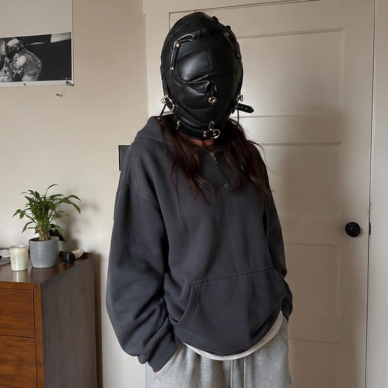 May include: A black leather-like full-head mask featuring multiple straps, metal buckles, and circular hardware accents. The mask is worn with a dark charcoal gray quarter-zip hooded sweatshirt and light gray sweatpants, creating a layered look.