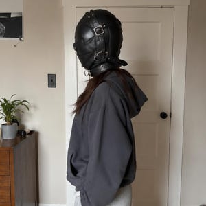 May include: A black leather head covering featuring a quilted texture, multiple adjustable silver-toned metal buckles, and a wide collar strap with metal hardware. The headgear is worn with a plain dark gray hooded sweatshirt and light gray pants.