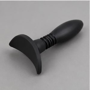 Könnte beinhalten: Ein mattschwarzes Handwerkzeug mit einem glatten, länglichen und abgerundeten Griff, der mit einem flexiblen, gerippten Halsbereich und einer breiten, geschwungenen, halbmondförmigen Basis verbunden ist, die einen stabilen Halt bietet.