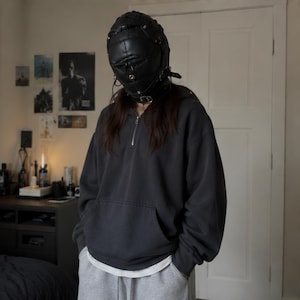 May include: A black leather-like full-head mask featuring multiple straps, buckles, and metallic hardware, worn with a dark charcoal gray quarter-zip hooded sweatshirt, a white undershirt, and light gray sweatpants with visible pockets.