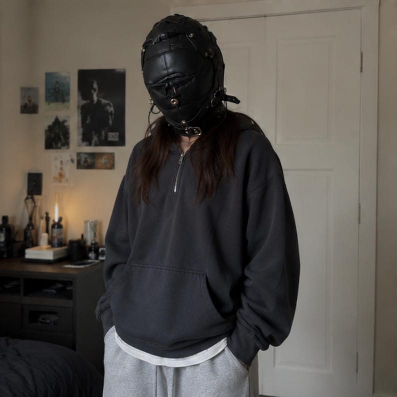 May include: A black leather-like full-head mask featuring multiple straps, buckles, and metallic hardware, worn with a dark charcoal gray quarter-zip hooded sweatshirt, a white undershirt, and light gray sweatpants with visible pockets.