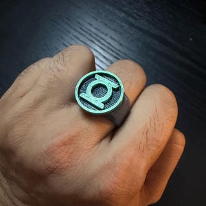 Puede incluir: Un anillo con una cara circular que presenta un emblema en relieve de color verde metálico del logotipo de Green Lantern sobre un fondo oscuro y texturizado. La banda del anillo es de un material gris oscuro mate con un acabado de capas impresas en 3D.