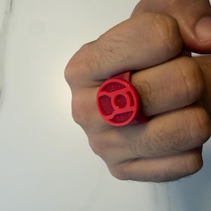 Puede incluir: Un anillo de color rojo brillante que presenta una cara circular con un emblema en relieve. El anillo tiene un acabado mate y texturizado, consistente con una construcción de plástico impreso en 3D, mostrando un color sólido y definido.