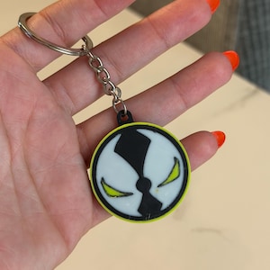 Puede incluir: Un llavero circular con un diseño gráfico en blanco y negro de un rostro estilizado con ojos afilados de color verde lima. El emblema está bordeado por un fino borde verde lima y está unido a una anilla de metal plateado mediante una cadena.