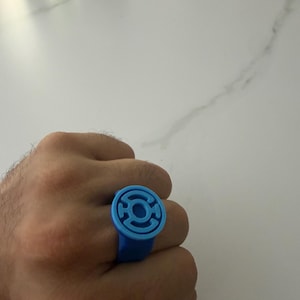 Peut inclure: Une bague bleu clair présentant une face circulaire avec un emblème en relief au design stylisé. La bague possède une finition mate et lisse ainsi qu'un anneau solide qui entoure le doigt, affichant une teinte monochrome sur l'ensemble.