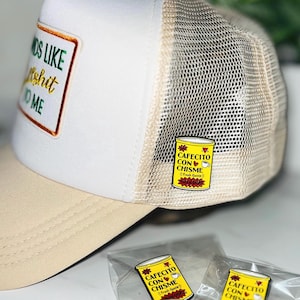 May include: A trucker hat with a white front panel, beige brim, and mesh back. The hat has a patch with the text "OS LIKE Ashit ME". A yellow pin with the text "CAFECITO CON CHISME" is attached to the hat. Additional pins are visible.