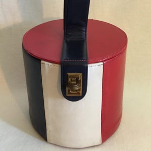 May include: A cylindrical handbag featuring vertical panels in navy blue, white, and red. The bag has a matching navy blue top handle and a central navy blue strap secured with a gold-toned metal latch closure on the front of the cylindrical body.