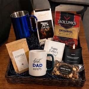 Op de afbeelding: Een geschenkmand met een blauwe mok met karabijnhaak, een witte mok met de tekst Best Dad Ever, een Lindt 70% cacao chocoladereep, een zak Jack Link's beef jerky, een zak koffie, een Happy Father's Day kaart, een brandende kaars en gemengde noten.