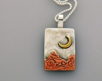 May include: A rectangular pendant with a silver chain. The pendant features a textured silver background with a gold crescent moon and copper-colored mountain range design. The chain is a silver box chain.