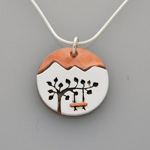May include: A silver necklace with a round pendant featuring a copper-colored mountain scene with a tree and a swing. The pendant is made of silver and copper.