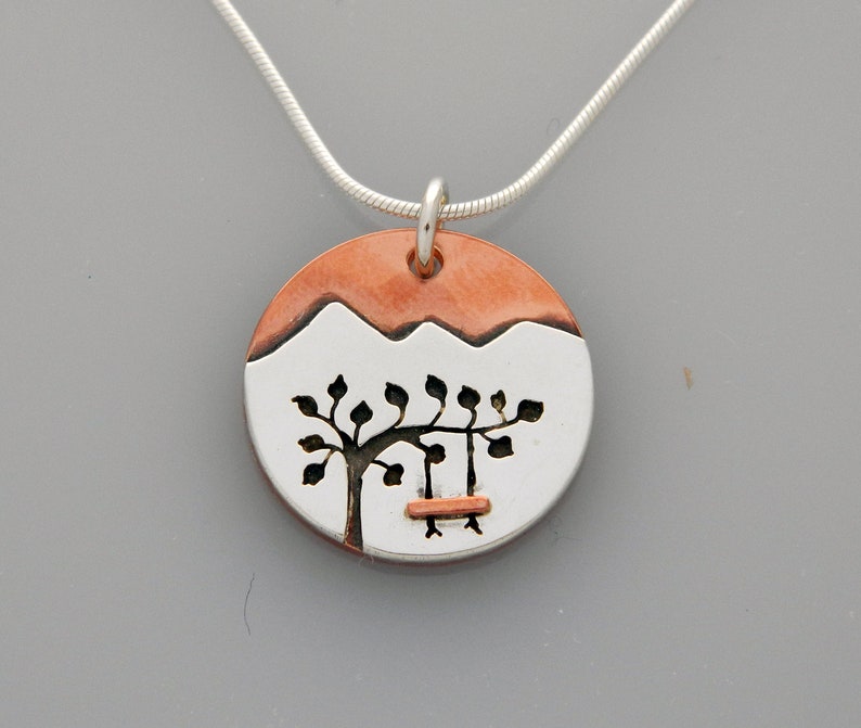 May include: A silver necklace with a round pendant featuring a copper-colored mountain scene with a tree and a swing. The pendant is made of silver and copper.