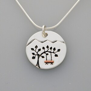 May include: Silver necklace with a round pendant featuring a stylized tree with a swing hanging from its branches. The pendant is textured and has a mountain range design at the top. The swing is made of copper.