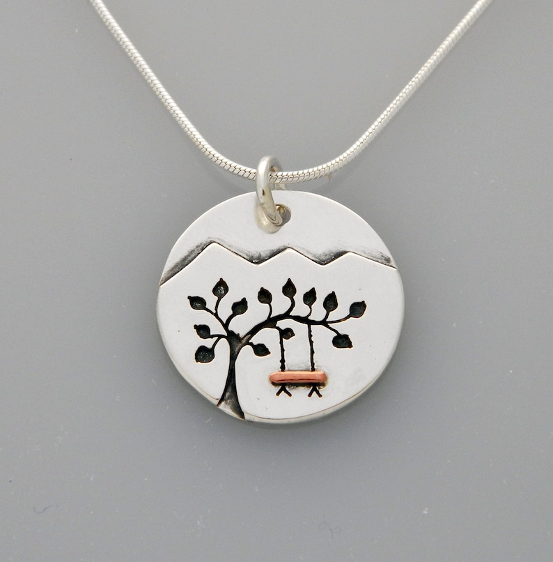 May include: Silver necklace with a round pendant featuring a stylized tree with a swing hanging from its branches. The pendant is textured and has a mountain range design at the top. The swing is made of copper.