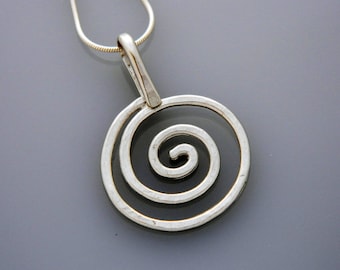 Puede incluir: Un collar de plata con un colgante en espiral. El colgante está hecho de dos círculos concéntricos, con el círculo interior formando una espiral.