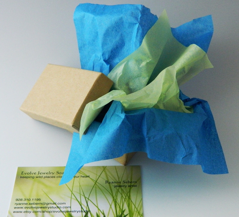 May include: A brown cardboard box wrapped in blue and green tissue paper. The box is labeled "Evolve Jewelry Studio" and includes contact information. The text "keeping wild places close to our heart" is also visible on the box.