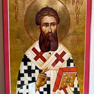 Könnte beinhalten: Ein gerahmtes religiöses Bildnis einer bärtigen Figur mit Heiligenschein vor goldenem Hintergrund. Die Figur trägt ein weißes Gewand mit schwarzem Kreuzmuster und eine weiße Stola mit roten Kreuzen und hält ein rotes Buch mit Gold.