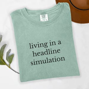 Puede incluir: Camiseta verde salvia Comfort Colors con la frase "living in a headline simulation" impresa en negro. La camiseta está doblada y sobre una superficie blanca, con un sombrero marrón visible al fondo.