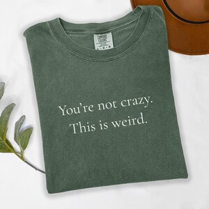May include: A sage green t-shirt with the text "You're not crazy. This is weird." in white. The shirt has a crew neck and short sleeves. The Comfort Colors brand label is visible on the upper back of the shirt.