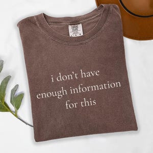 May include: A brown Comfort Colors t-shirt with the text "i don't have enough information for this" in white. The shirt is folded and has a crew neck. A brown hat is partially visible in the background.