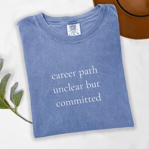以下が含まれることがあります： 白文字で「career path unclear but committed」と書かれた青色のComfort Colors Tシャツ。シャツは水色で、クルーネックです。背景には茶色の帽子が一部見えています。
