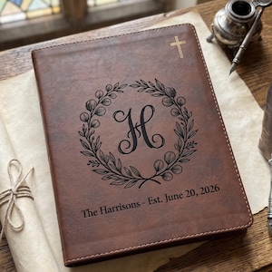 May include: A brown leather-like journal featuring a gold cross at the top, a central black engraved wreath surrounding the letter H, and the text The Harrisons - Est. June 20, 2026, with visible stitching along the edges of the cover.