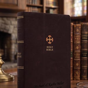May include: A dark brown leather-bound Holy Bible featuring gold foil stamping on the cover and spine. The cover displays a geometric emblem, the text Holy Bible, and the personalized inscription Michael & Kathy Miller Est. August 21st, 2026.