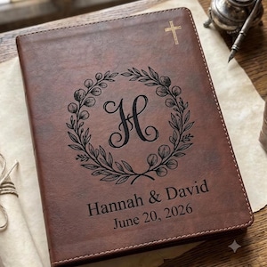 May include: A brown leather-like journal featuring a gold-colored cross at the top. The center displays a black engraved floral wreath surrounding the letter H. Below the wreath, the text reads Hannah & David and the date June 20, 2026.