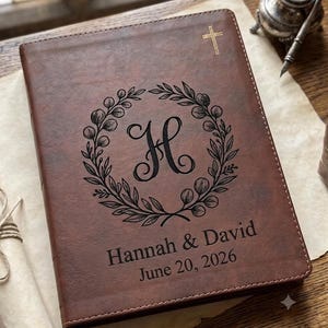 May include: A brown leather-like journal featuring a gold-colored cross at the top. The center displays a black engraved floral wreath surrounding the letter H. Below the wreath, the text reads Hannah & David and the date June 20, 2026.