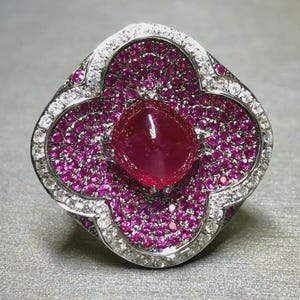 May include: An elaborate ring with a central oval ruby, encircled by smaller pink gemstones. The ring's design is a four-leaf clover shape, edged with white gemstones. The ring is displayed on a grey surface.