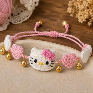 May include: A handmade crochet bracelet featuring a white cat face with a pink bow, flanked by pink crocheted hearts and white crocheted accents. The bracelet includes small gold-toned metal bells and has an adjustable pink cord closure.
