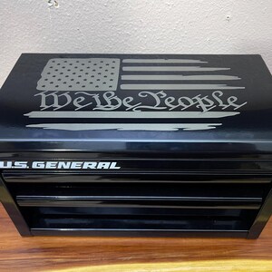 May include: Black metal tool chest with three drawers, featuring the text "U.S. GENERAL" and an American flag design with the words "We the People" on the lid. The chest is on a wooden surface.