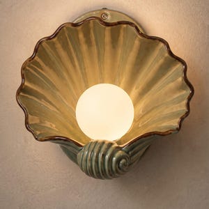 May include: A wall-mounted light fitting shaped like a large, open seashell with a glossy, pale green finish and dark brown edges. A single, round, frosted white light bulb is positioned in the centre of the shell, emitting a warm, soft glow.