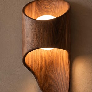 Può includere: Una lampada da parete realizzata in legno marrone scuro con una forma curva e organica. L'apparecchio presenta una lampadina centrale in vetro bianco che emette una luce calda, illuminando le venature del legno e proiettando luce verso l'alto.