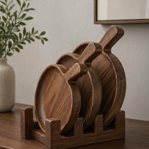 Könnte beinhalten: Ein Set aus vier Holzservierplatten mit Griffen, präsentiert in einem passenden Holzhalter. Die Platten sind rund mit einer dunkelbraunen Oberfläche, die die natürliche Holzmaserung zeigt. Der Halter hält die Platten aufrecht.