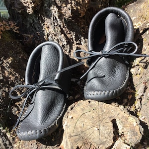 May include: A pair of black leather moccasins with a soft, textured appearance. The shoes have a rounded toe and are laced with a leather tie. The moccasins are displayed on a tree trunk.