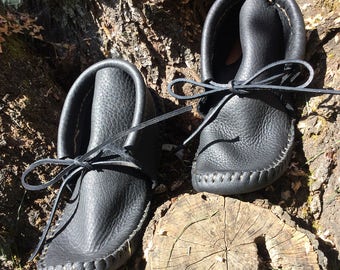 Puede incluir: Un par de mocasines de cuero negro con un aspecto suave y texturizado. Los zapatos tienen una punta redondeada y están atados con un lazo de cuero. Los mocasines se exhiben sobre un tocón de árbol.
