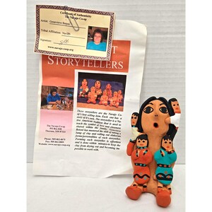 May include: A terracotta clay storyteller figurine featuring a central adult figure with an open mouth, surrounded by four smaller figures attached to the body. The figures are painted with orange, teal, and black details. Includes a certificate of authenticity.