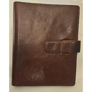 May include: A dark brown leather-like notebook cover with a smooth, glossy finish and a visible grain texture. The rectangular cover features a horizontal strap closure on the right side that secures the front flap to the main body of the item.