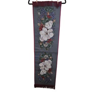 May include: A long, rectangular grey table runner featuring a woven design of white magnolia flowers, purple grapes, and green leaves. The runner has a deep burgundy border and matching burgundy fringe at both the top and bottom edges.