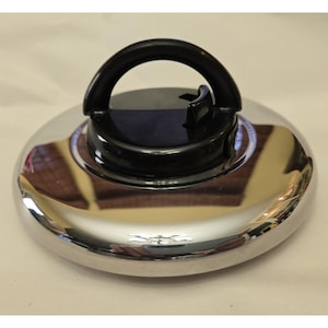 Puede incluir: Una tapa circular de cromo pulido para olla con un asa central de plástico negro. El asa es arqueada e incluye una pequeña perilla de ajuste negra integrada en el lateral. La superficie metálica es reflectante y tiene un borde liso.