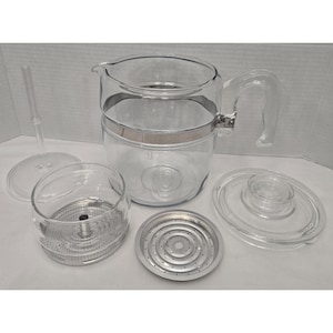 May include: A clear glass coffee percolator set featuring a main carafe with a handle and metal band, a glass lid, a perforated metal basket, a central stem, and a circular metal base plate. All components are disassembled and arranged together.