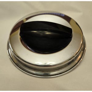 Puede incluir: Una tapa circular para olla hecha de acero inoxidable pulido con una superficie lisa y reflectante, que presenta un asa arqueada de plástico negro en el centro. La tapa tiene una forma ligeramente abombada con un borde elevado alrededor.