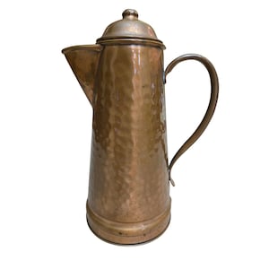 Può includere: Una caffettiera vintage in rame con una texture martellata. La caffettiera presenta un beccuccio, un manico curvo e un coperchio con un piccolo pomello. Il rame ha una calda tonalità marrone-rossastra.
